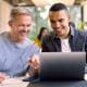 two men manager employee colleagues smiling looking at a laptop on a desk in an open office