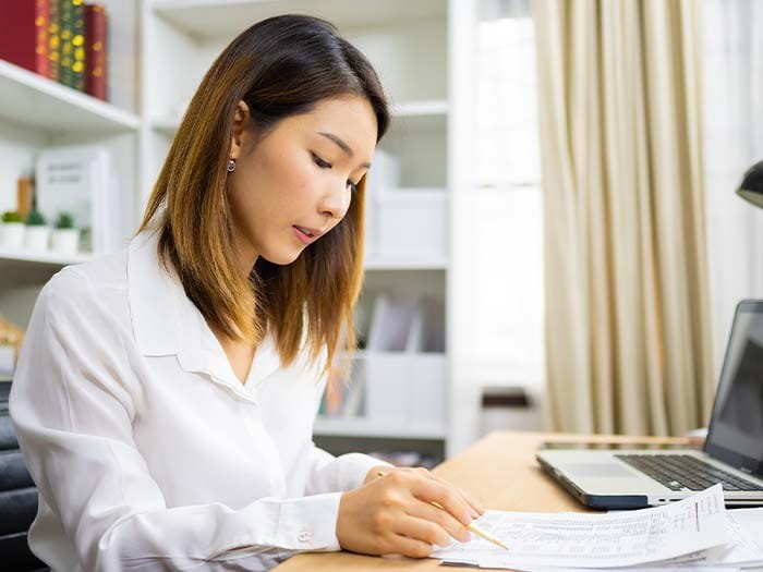 young asian woman working at a home office desk laptop computer writing with a pen filling out a form